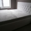 Čalouněná postel na zakázku je potažena bílou koženou. Čelo je vysoké a jeho potah je ozdoben velkými knoflíky a sešíváním do kosočtverců. 